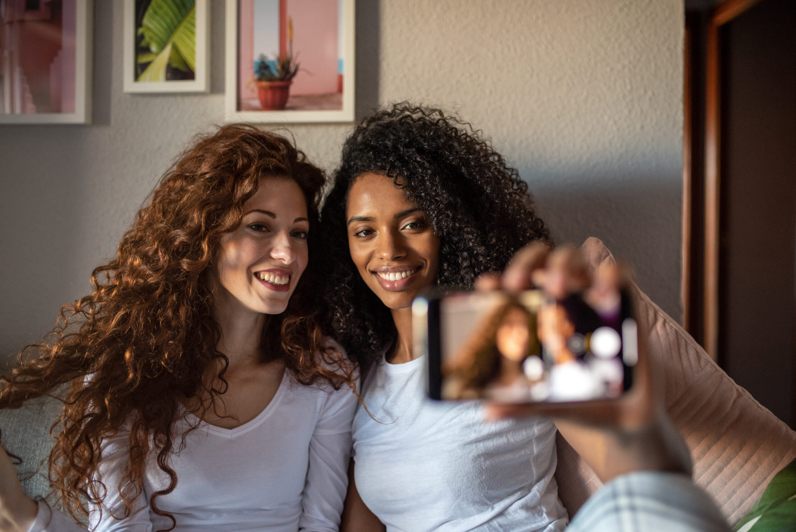 Young cheerful women friends seating relaxed on the sofa with mobile phone taking a selfie